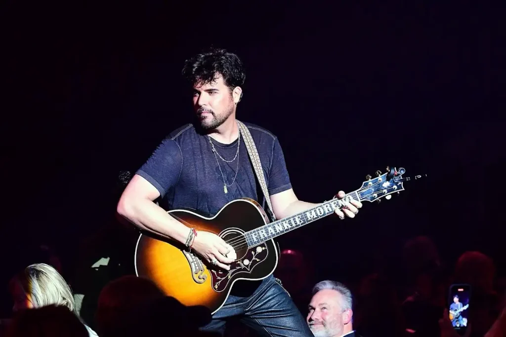 A male musician with a beard plays an acoustic guitar with "FRANKIE MORENO" written on the neck.