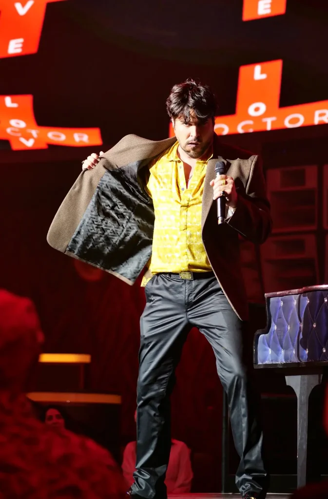 A man in a yellow shirt, brown jacket, and black pants holds a microphone, with "LOVE DOCTOR" on red signs.