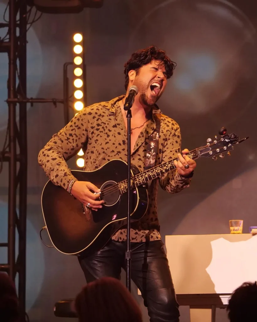 A man with dark curly hair, wearing a leopard print shirt and black leather pants, sings into a microphone while playing an acoustic guitar on a stage.