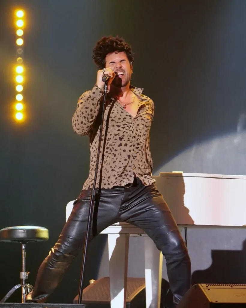 A man with dark curly hair, wearing a leopard print shirt and black leather pants, sings into a microphone.