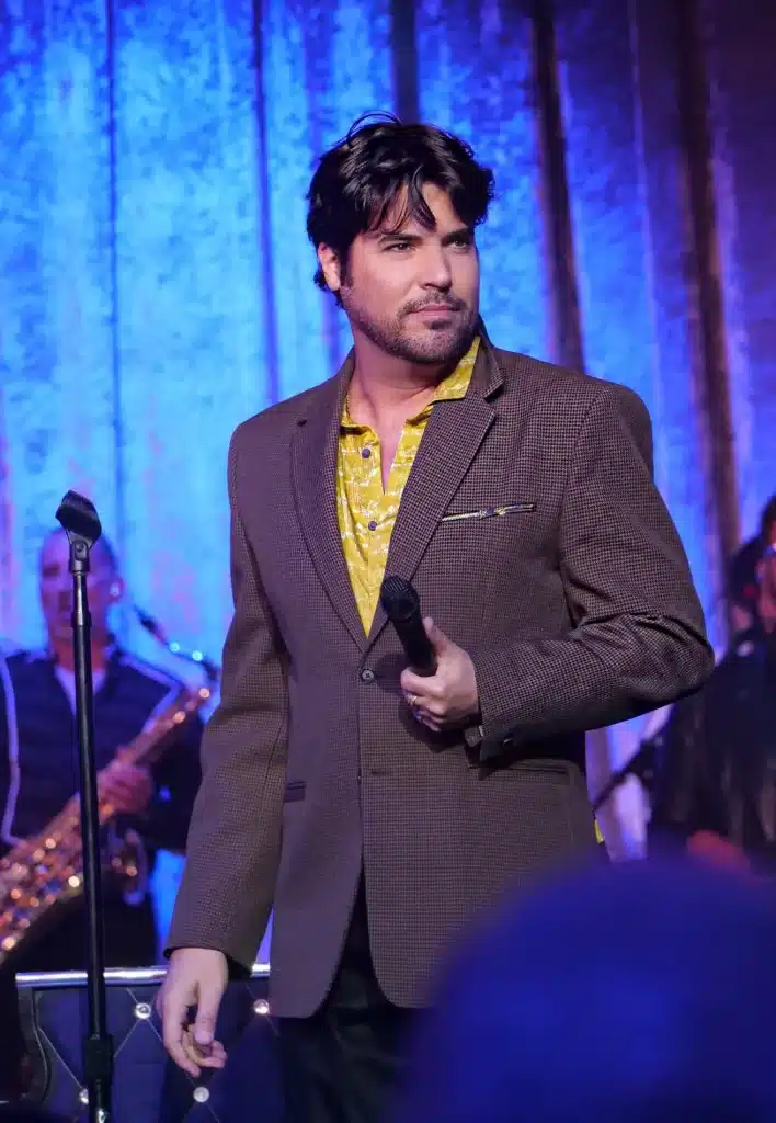 A man with dark hair and a beard, wearing a brown suit jacket and yellow shirt, holds a microphone on a blue-lit stage.