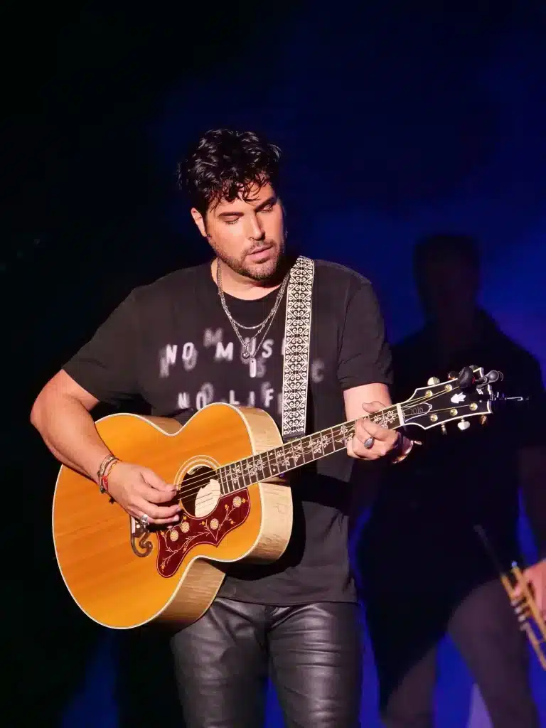 A man with dark hair and a beard plays an acoustic guitar, wearing a black shirt with "NO MUSIC NO LIFE" and leather pants.
