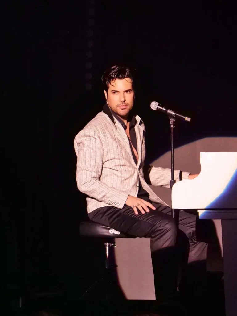 A man with dark hair and a striped jacket sits on a piano bench, looking at the viewer.