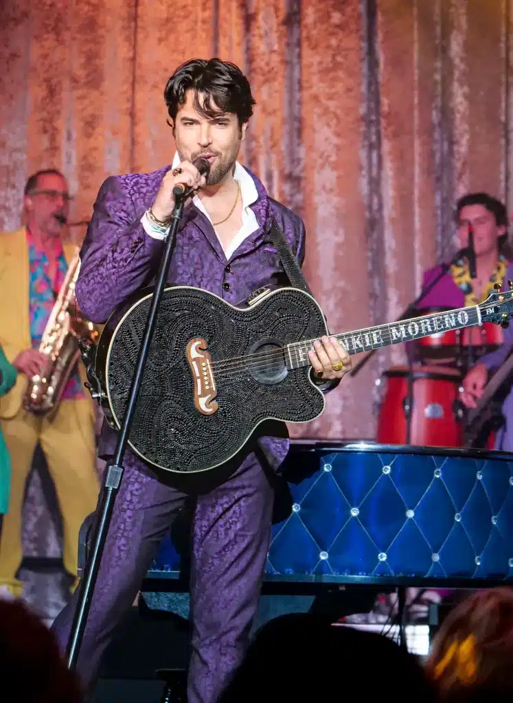 A man in a purple suit sings into a microphone while holding a black guitar with "FRANKIE MORENO" on the fretboard.