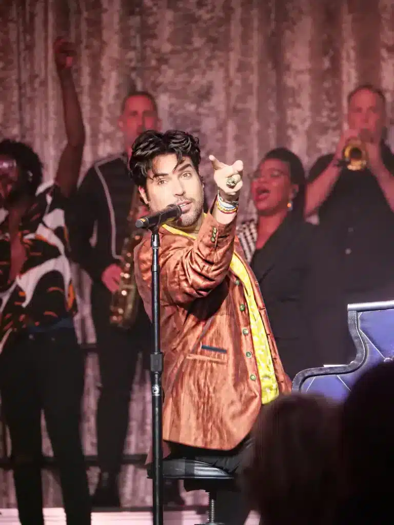 A male singer in a brown jacket and yellow shirt points to the right, surrounded by musicians on stage.