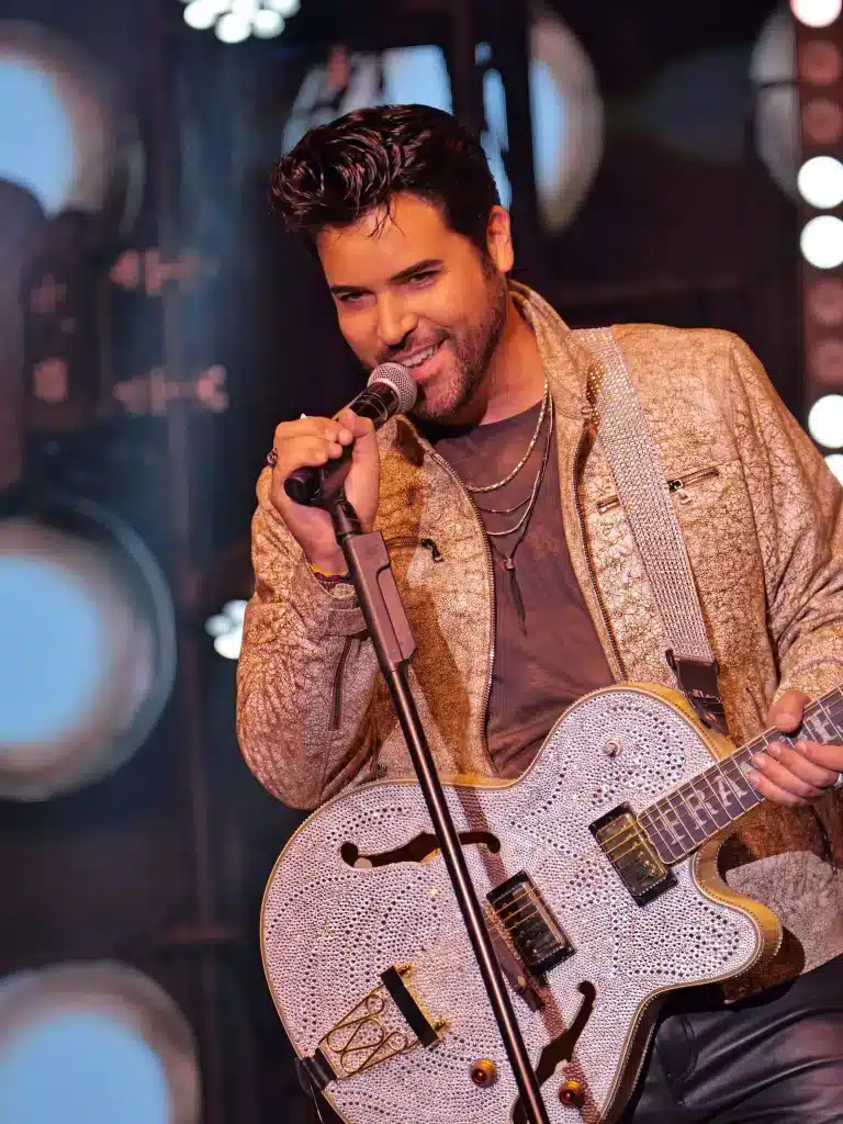 A smiling man with dark hair sings into a microphone, wearing a gold jacket and holding a sparkly guitar.