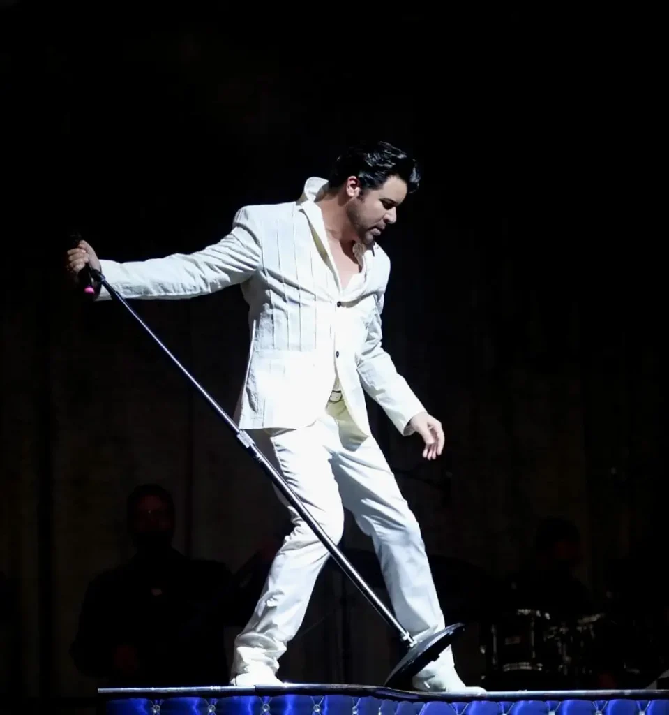 A man in a white suit, with dark hair, leans over a microphone stand on a dark stage.