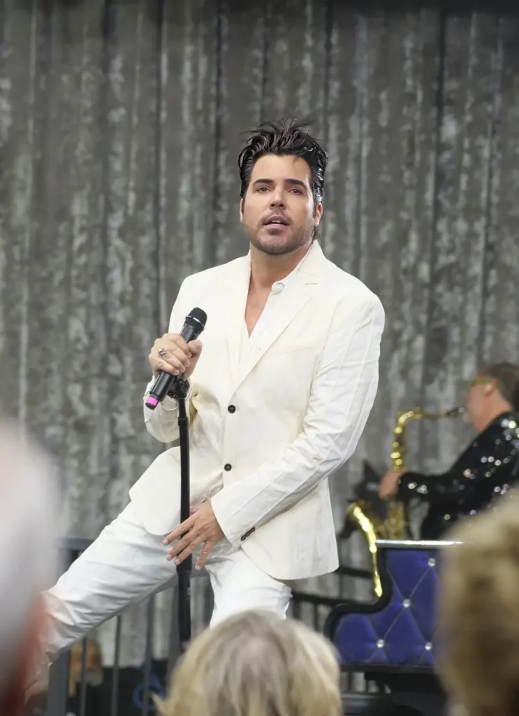 A man in a white suit holds a microphone, singing on stage with a saxophone player in the background.