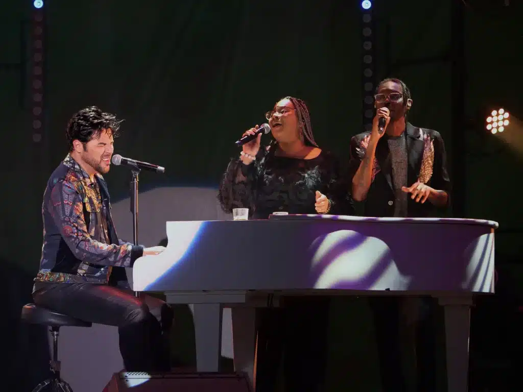 A man with a colorful jacket plays a white piano and sings into a microphone, with two backup singers.