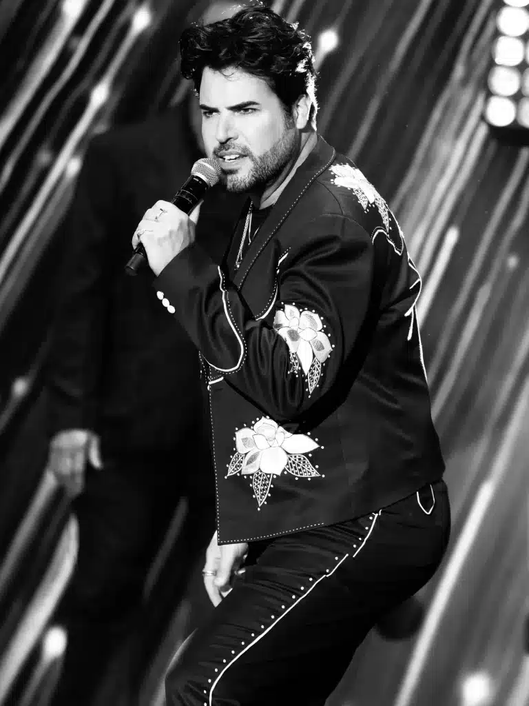 A man with dark curly hair and a beard sings into a microphone, wearing a black suit with white floral embroidery.