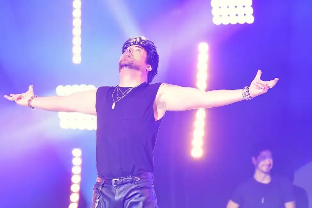 A man with dark hair and a beard, wearing a black sleeveless shirt, stands with arms outstretched, looking up at bright stage lights.