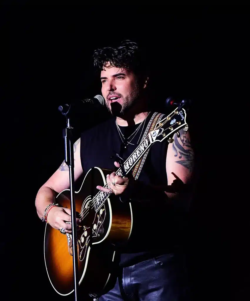 A man with dark hair and a beard sings into a microphone while playing an acoustic guitar with a "MORENO" strap.