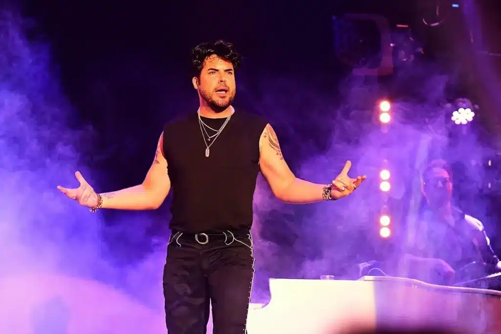 A male singer with dark hair and a black sleeveless shirt performs on stage with purple lights and smoke.