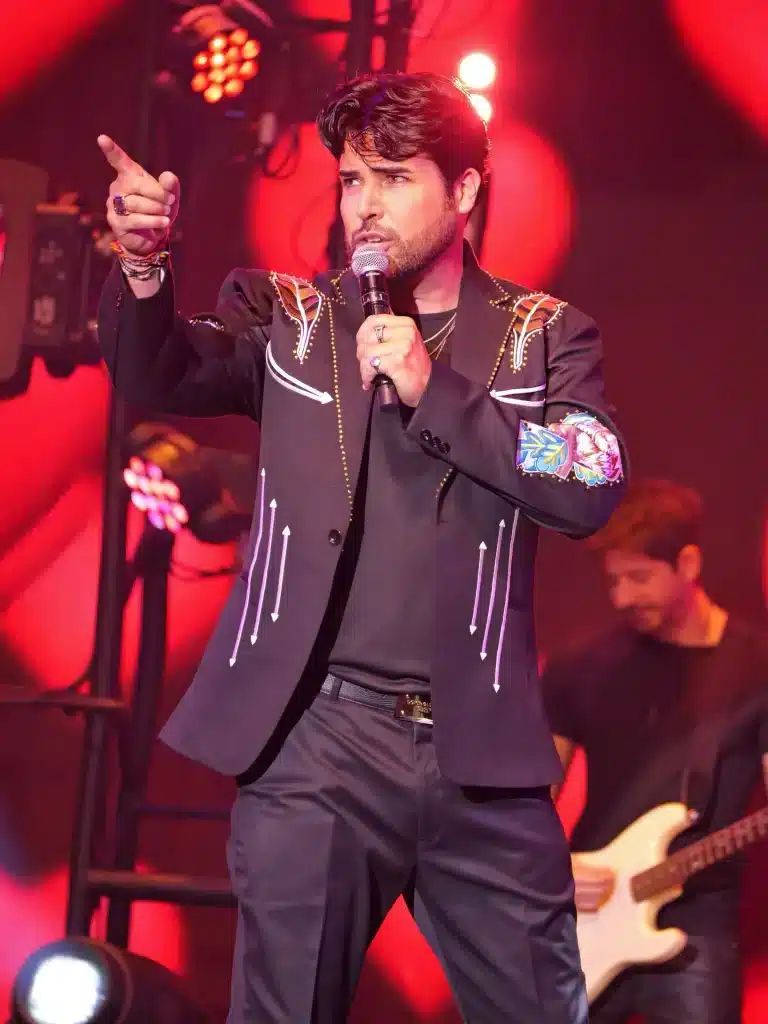 A man in a black western jacket with white embroidery sings into a microphone, pointing with his left hand.