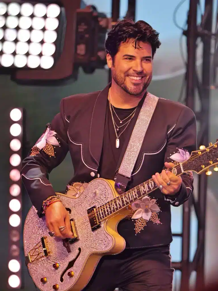 A smiling man with dark hair and a beard plays a rhinestone-covered guitar, wearing a black embroidered jacket.