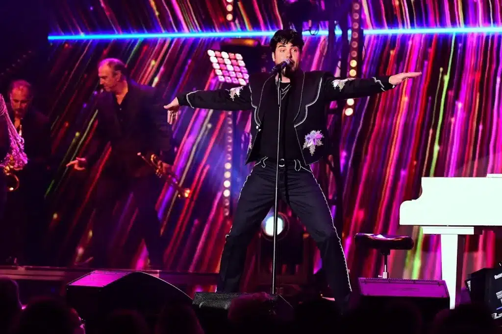 A male singer in a black embroidered jacket stands center stage with arms outstretched, backed by a band and colorful lights.