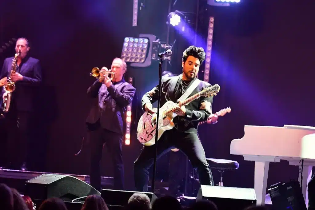 A band performs on stage with a guitarist in the foreground, a trumpeter, and a saxophonist.