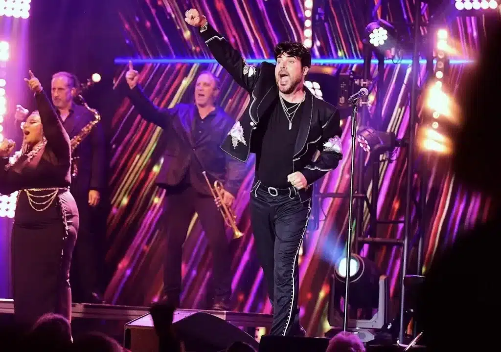 A male singer in a black embroidered jacket raises his fist on stage with backup singers and musicians.