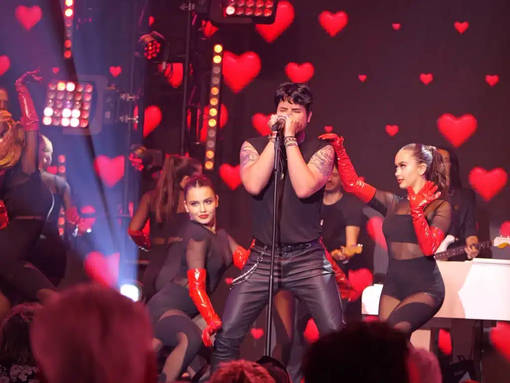 A male singer in leather pants performs on stage with female dancers in black outfits and red gloves.