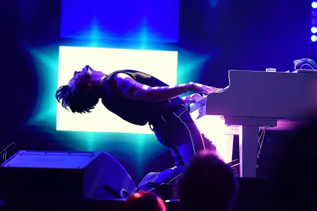 A male musician in a black tank top leans back dramatically while playing a white grand piano on a stage with blue and white lights.