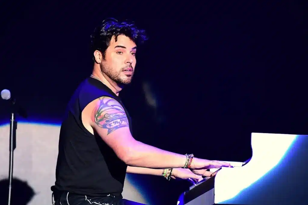 A man with dark hair and a beard, wearing a black sleeveless shirt, plays a white piano on stage.