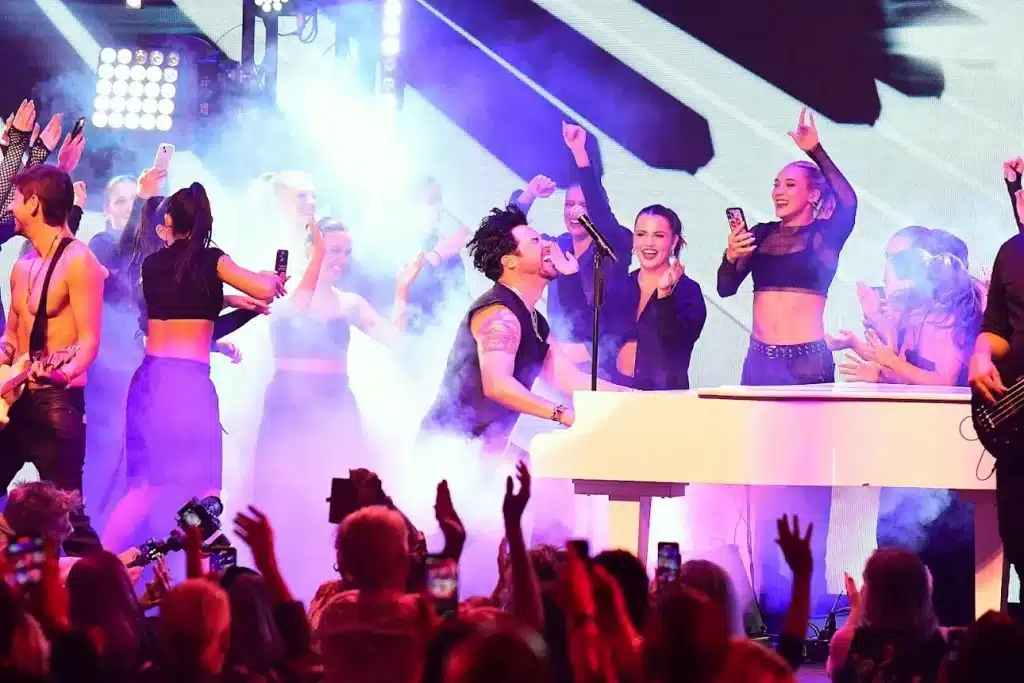 A band performs on stage with a pianist, guitarist, and singers, bathed in purple light and smoke.
