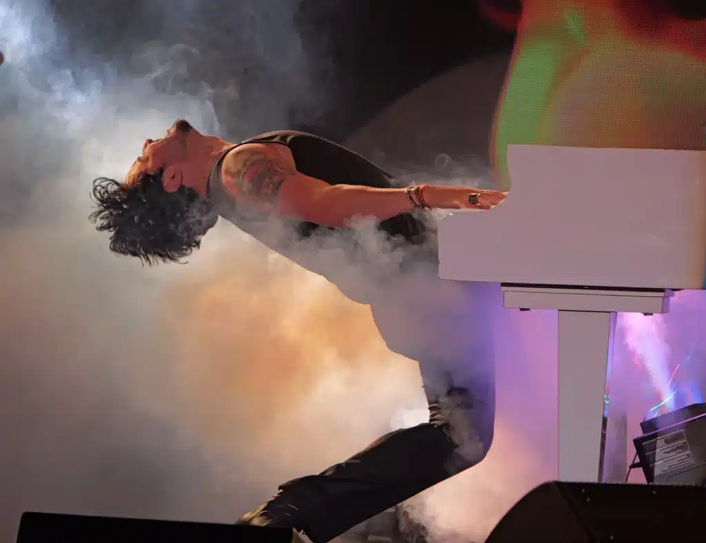 A male musician with tattoos on his left arm plays a white keyboard, leaning back into a cloud of smoke.