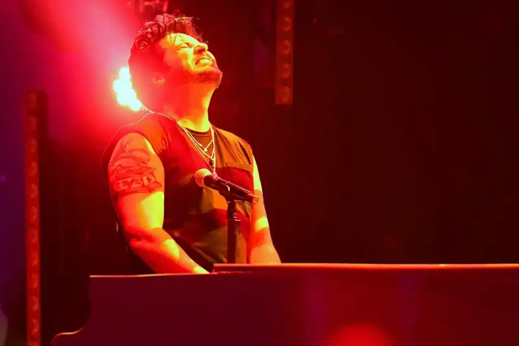 A man with a beard and tattoos on his left arm sings into a microphone under red stage lights.