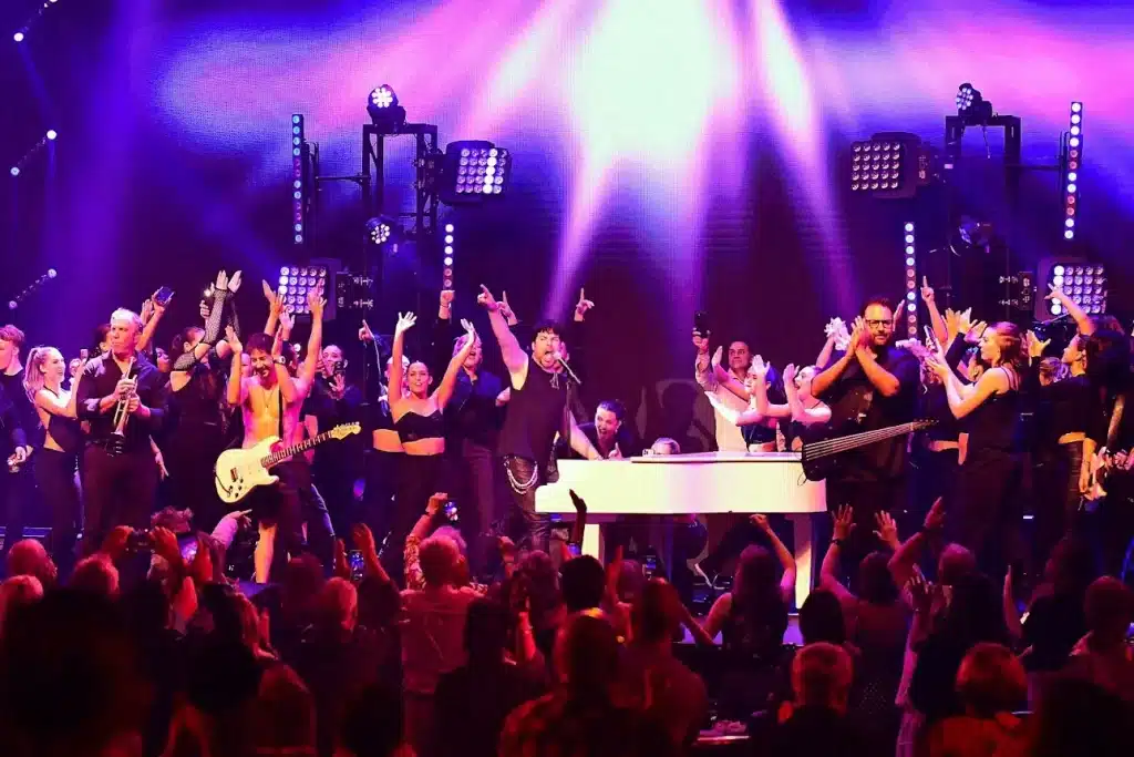 A large band and backup singers perform on stage under bright purple lights, with an audience in the foreground.