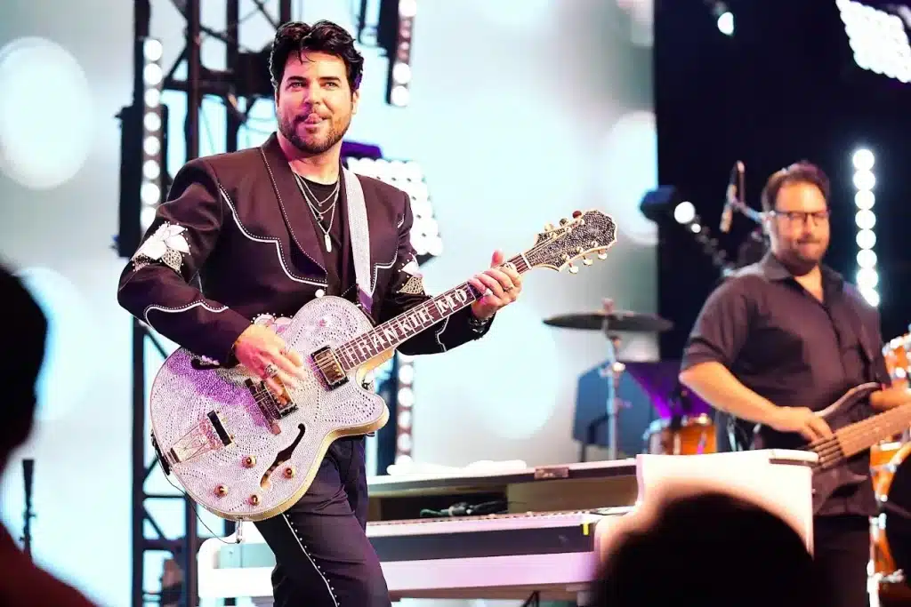 A man in a black western shirt with white embroidery plays a rhinestone-covered guitar that says "FRANKIE MORE."