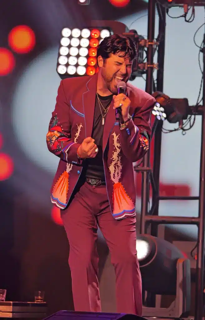 A man in a maroon suit with colorful embroidery sings into a microphone on a stage with bright lights.