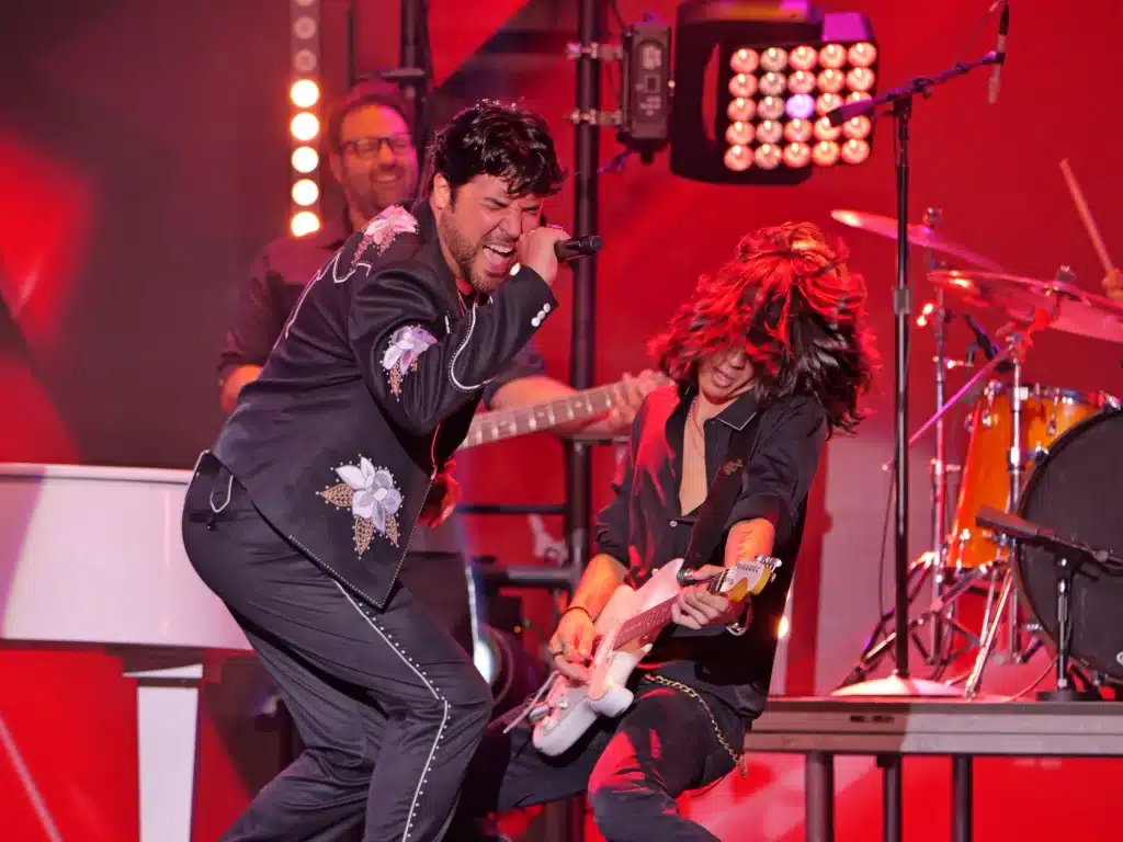 A singer in an embroidered black suit shouts into a microphone while a guitarist with long hair plays.