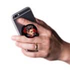 A hand holds a black smartphone with a pop socket featuring a man's head with dark hair and a red background.