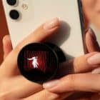 A person holds a white smartphone with a black pop socket featuring a man in a white suit and "FRANKIE" in red lights.