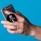 A hand holds a silver smartphone with a black pop socket featuring a white microphone and "FM" on a blue background.