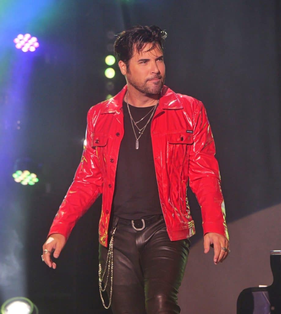 A man with dark hair and a beard wears a shiny red jacket, black shirt, and leather pants on stage.