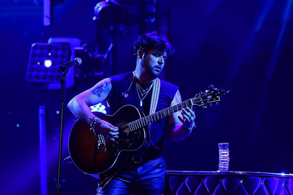 A male musician with tattoos and necklaces plays an acoustic guitar on a dark stage under blue light.