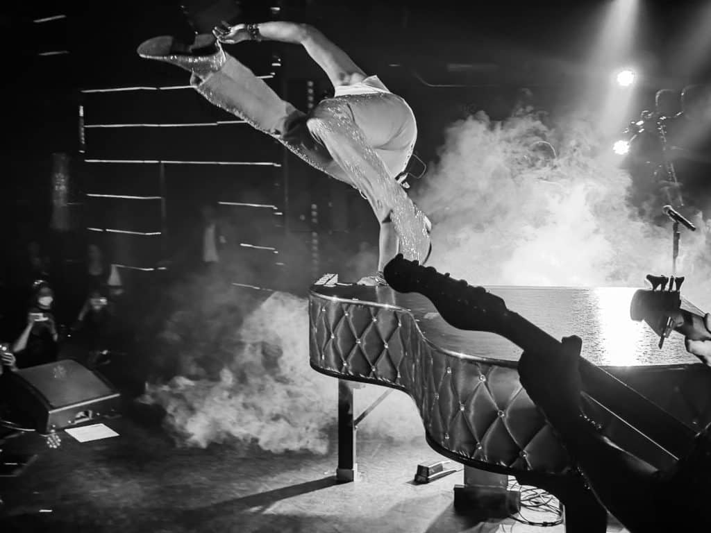 A performer in shiny pants and heels leaps over a quilted piano on a smoky stage, with guitars in the foreground.