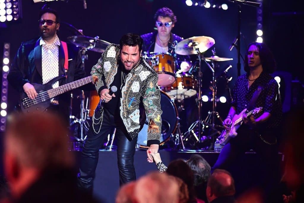 A male singer in a bedazzled jacket holds a microphone and reaches for a fan's hand on stage.