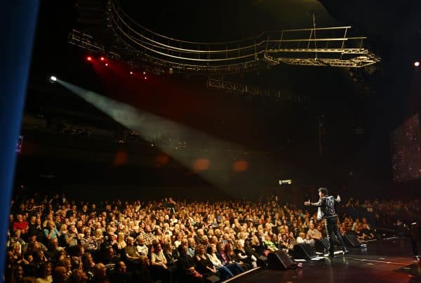 A performer in a black jacket with white designs stands on a stage, facing a large audience in a dark theater.